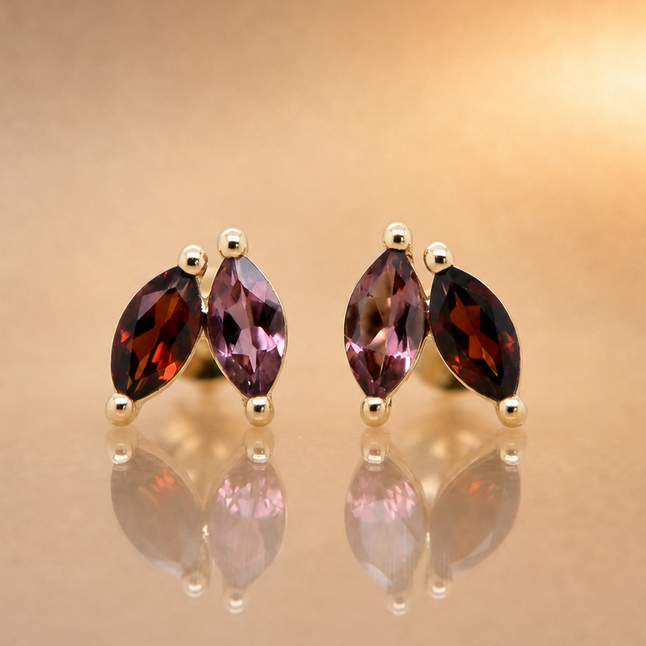 A pair of gold stud earrings featuring marquise-cut purple and dark red gemstones, displayed on a reflective gold surface against a warm beige background.