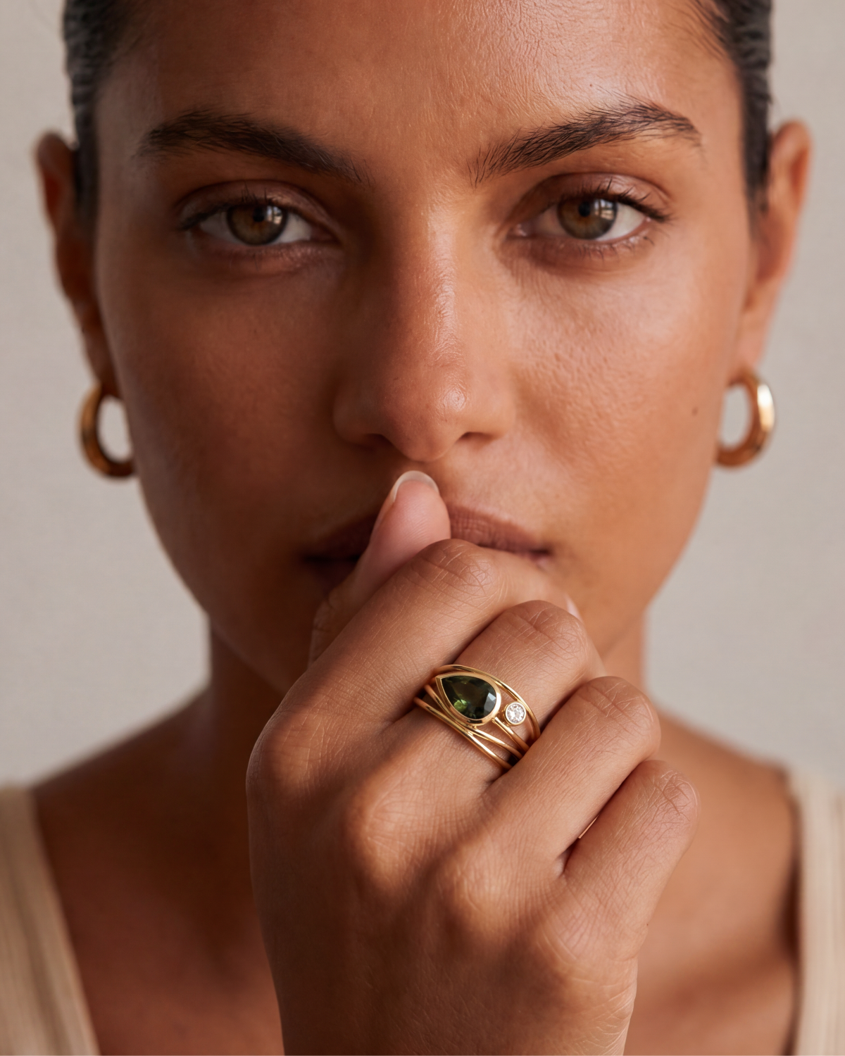 A close-up portrait of a woman wearing a gold ring with a green heart-shaped stone on her finger, along with gold hoop earrings, set against a neutral beige background.