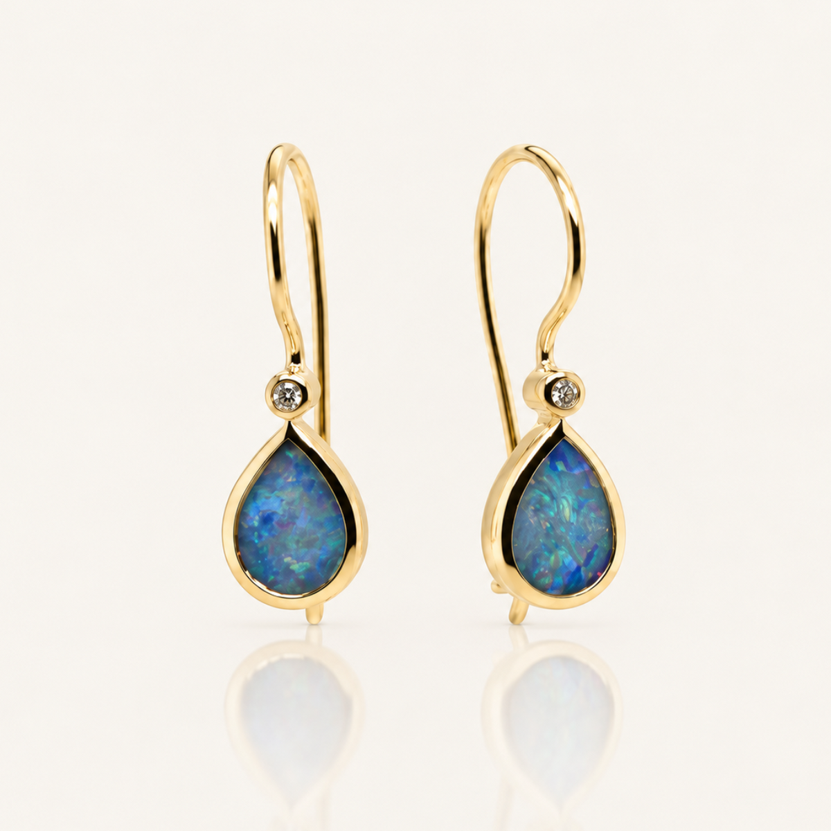 A pair of gold drop earrings with teardrop-shaped blue opal centers and a small clear stone above each opal, shown on a clean white background with a soft reflection below.
