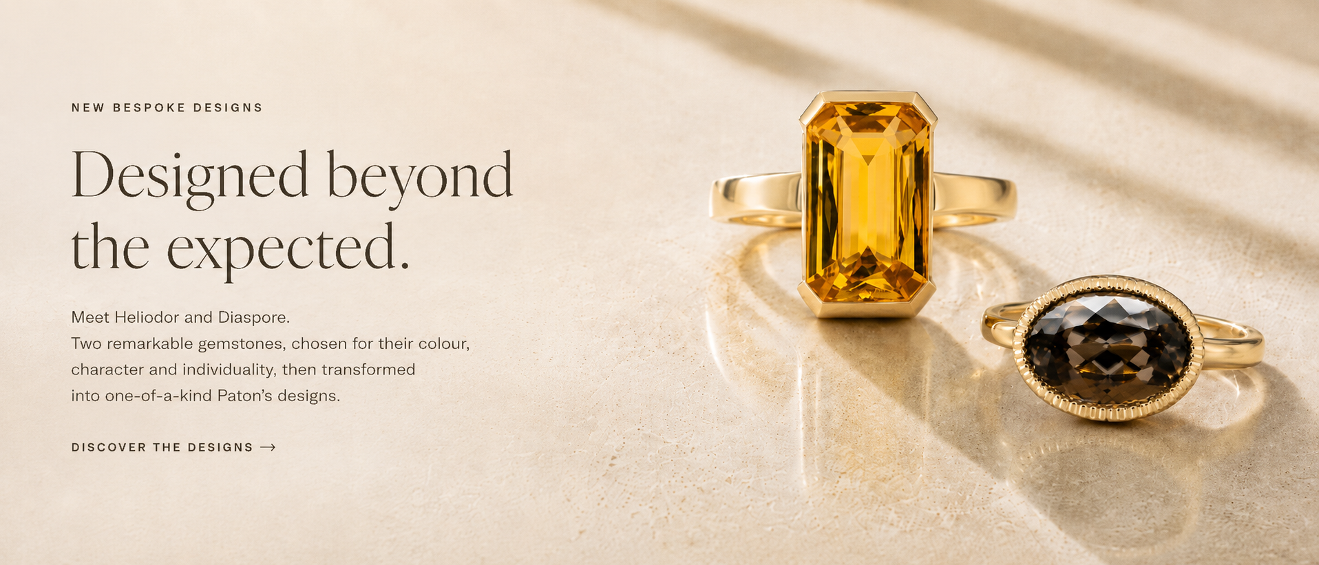 Two gold rings displayed on a light beige surface with elegant shadows. The larger ring features a rectangular yellow gemstone, and the smaller ring features an oval dark gray gemstone.