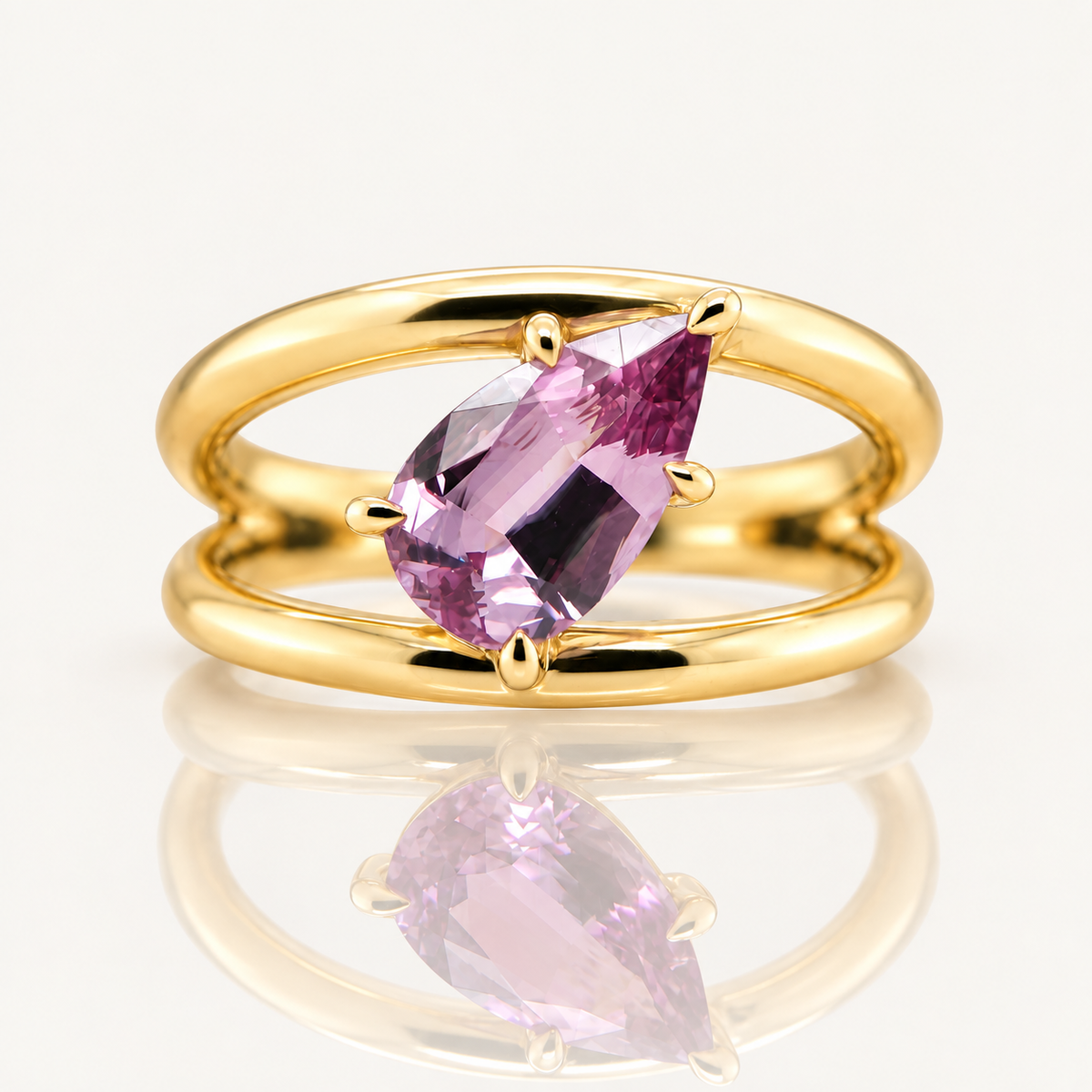 A gold ring with a pear-cut pink/purple gemstone in a prong setting, displayed on a clean white reflective surface.