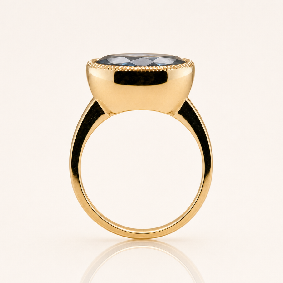 A gold ring with a large, polished oval cut gemstone in a bezel setting, shown on a clean off-white background with a soft reflection below.