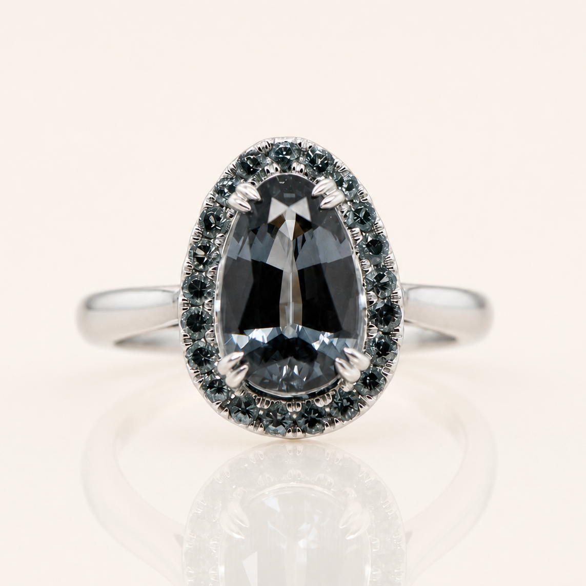 A white gold ring featuring a large pear-cut dark gray gemstone in a halo setting with smaller dark blue-gray stones, shown on a clean off-white background with a soft reflection below.
