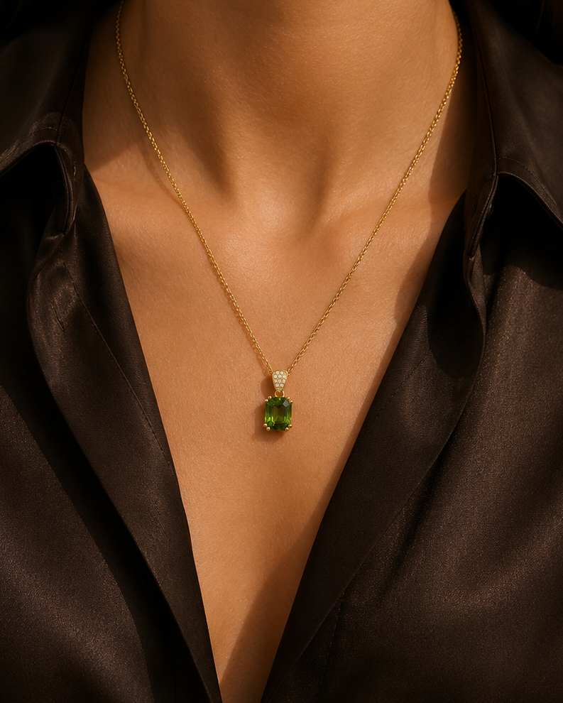 A close-up of a model wearing a delicate gold chain necklace with a green faceted pendant, styled with a dark brown satin shirt.